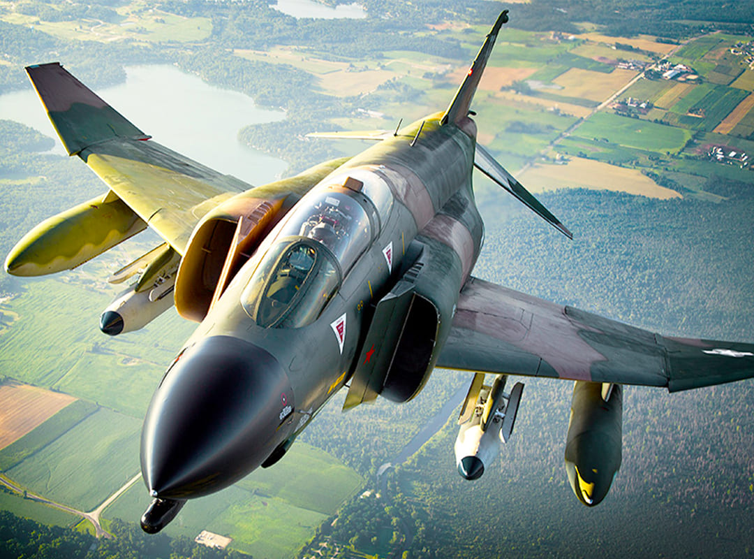 F-4D Phantom II military jet flying at altitude over a patchwork of fields and forests, viewed from above during flight.