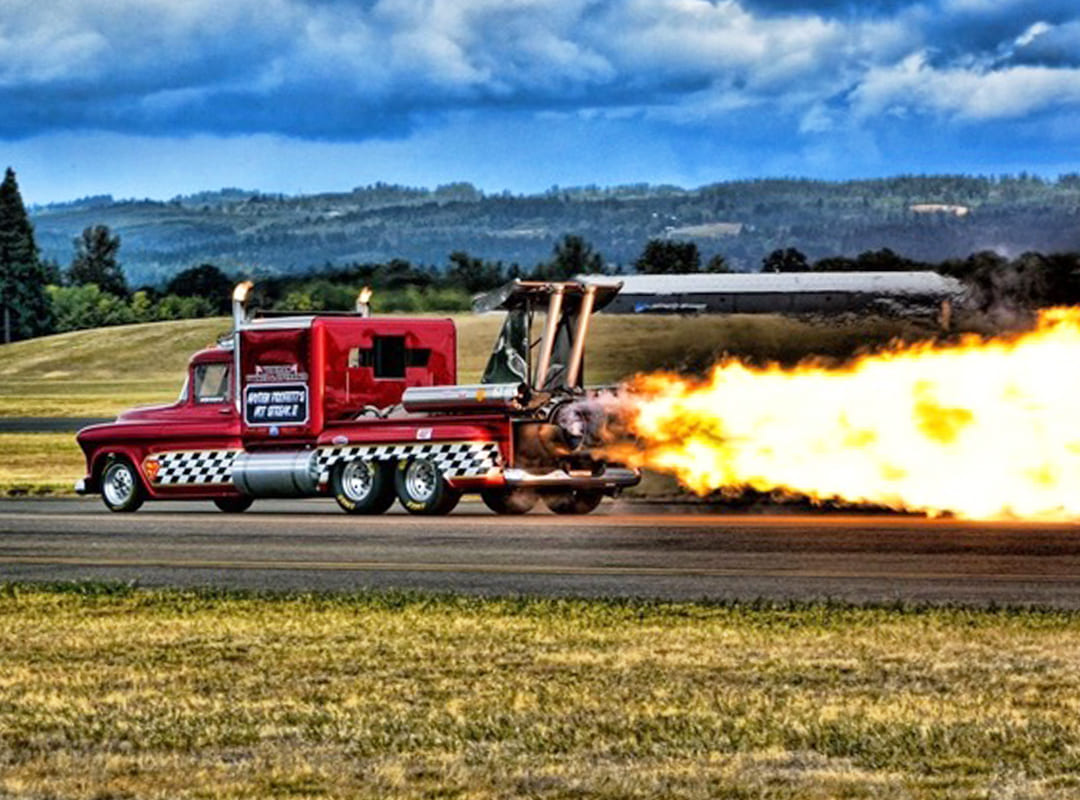 Smoke-n-Thunder Jet Truck racing down an airfield runway with a powerful jet engine producing a large flame plume during an airshow performance.