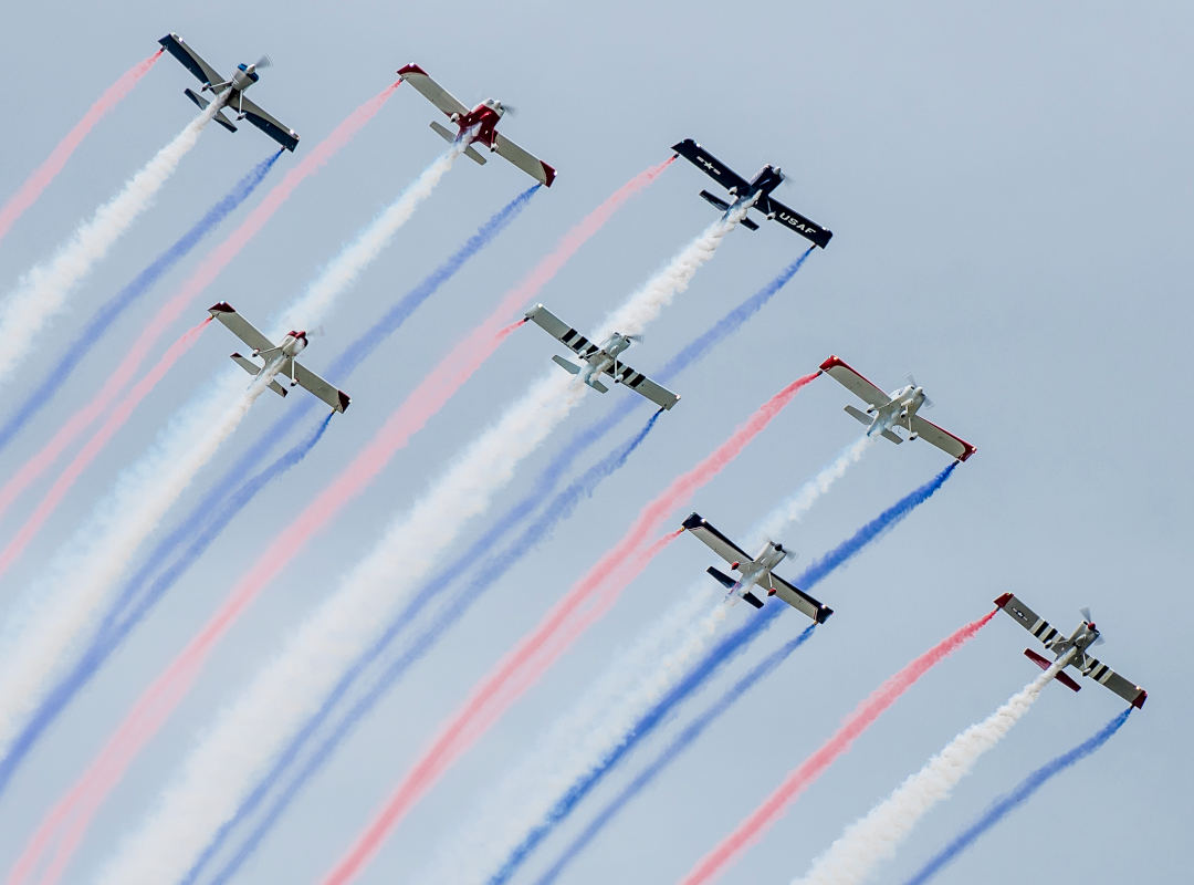KC Formation Flight Team flying in tight formation during an airshow, with multiple aircraft leaving red, white and blue smoke trails against the sky.