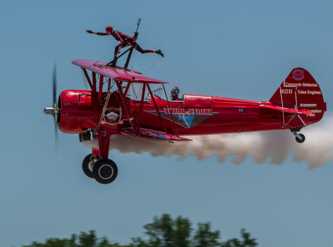 Third Strike Wingwalking Team performer standing on the wing of a red biplane in flight during an airshow, with smoke trailing behind the aircraft.