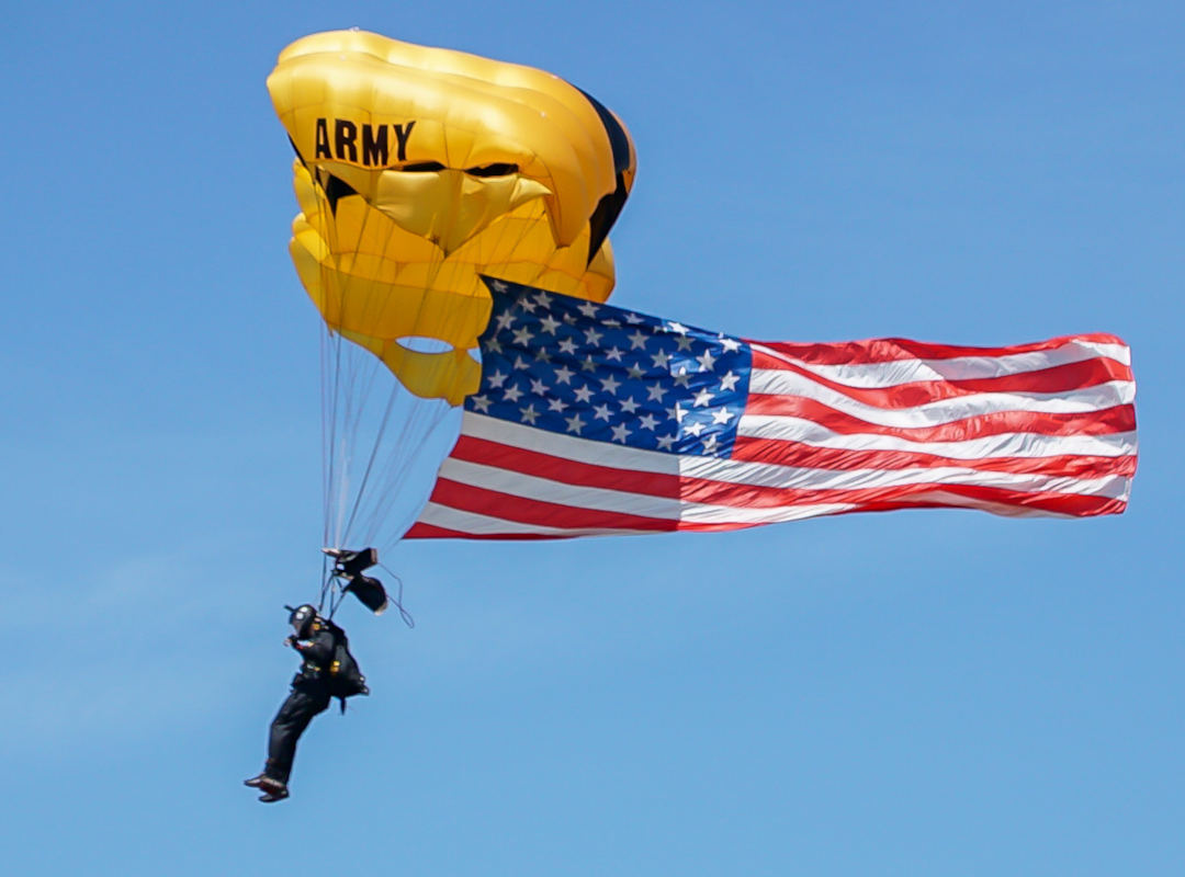 U.S. Army Golden Knights parachutist descending under a yellow canopy while carrying a large American flag against a clear blue sky.