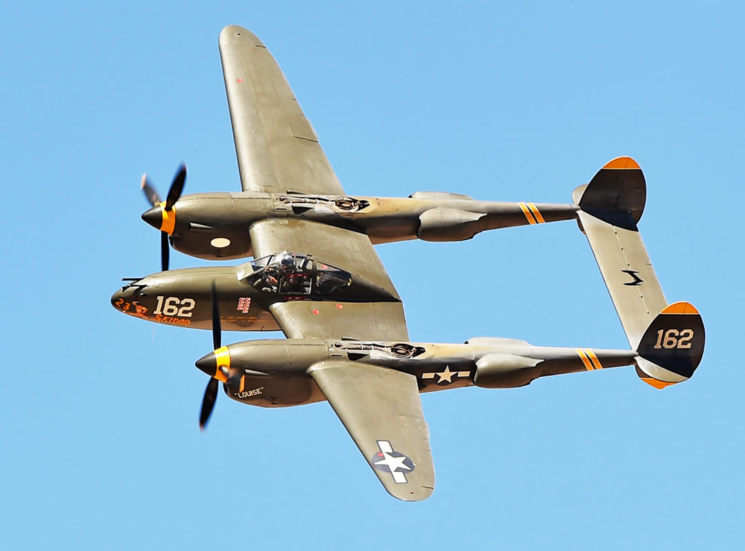 Alt Text: World War II P-38J Lightning twin-engine fighter aircraft in flight against a clear blue sky, featuring olive drab paint, twin tail booms, spinning propellers, U.S. Army Air Forces star insignia, and the number “162” displayed on the nose and tail.