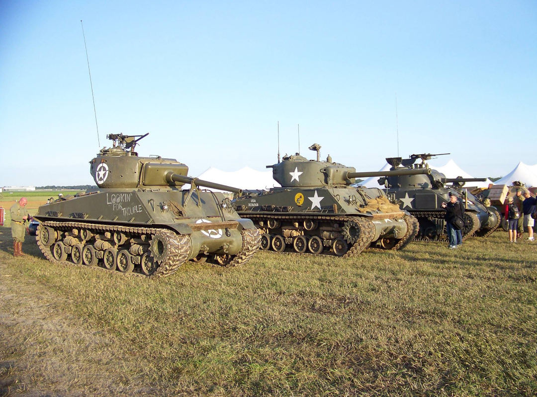 Three restored World War II M4 Sherman tanks, known collectively as the “Three Kings of Columbus,” parked side by side on grass at the Columbus, Ohio Air Show. The olive drab tanks display white U.S. Army star insignia and turret-mounted cannons, with event tents and spectators visible in the background under a clear sky