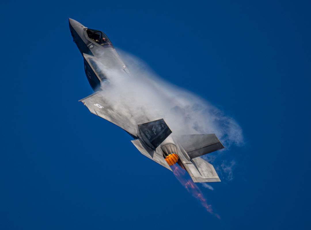U.S. Air Force F-35 Lightning II fighter jet climbing steeply with afterburner engaged, vapor clouds forming around the wings against a clear blue sky.